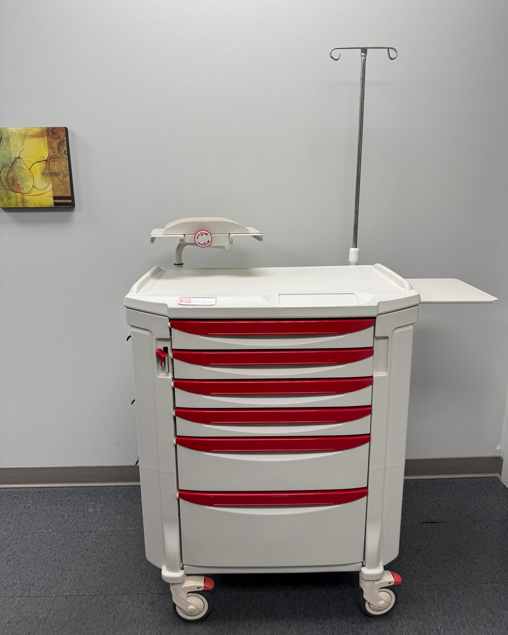 A white and red medical procedure cart with multiple drawers and a worksurface, equipped with a IV pole and lock system, set against a room with a wall painting.